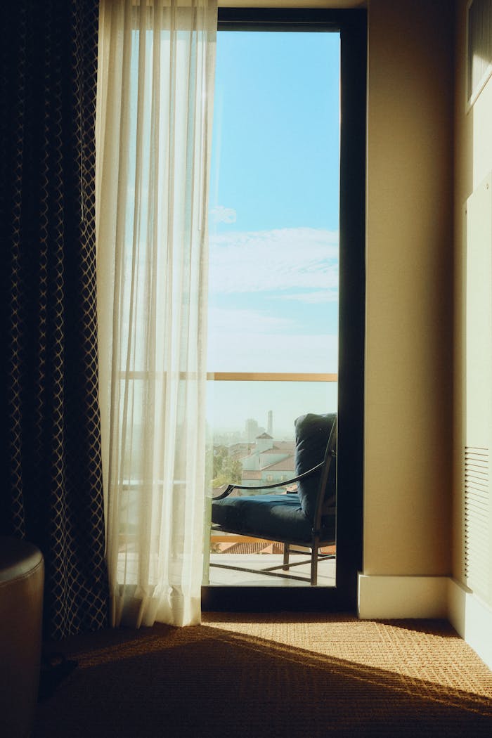 A serene hotel room with elegant sheer curtains opening to a sunlit balcony view.
